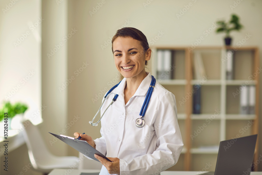 Young smiling doctor therapist looking at camera and making notes over medical clinic office at background Young smiling doctor therapist looking at camera and making notes over medical clinic office at background