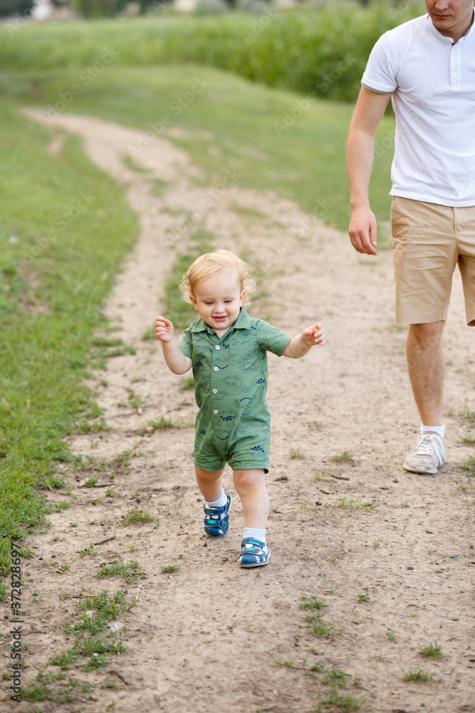 Fototapeta premium Cute little boy walking along the path on the background of reeds. His father is with him. Walk in park. Family life.