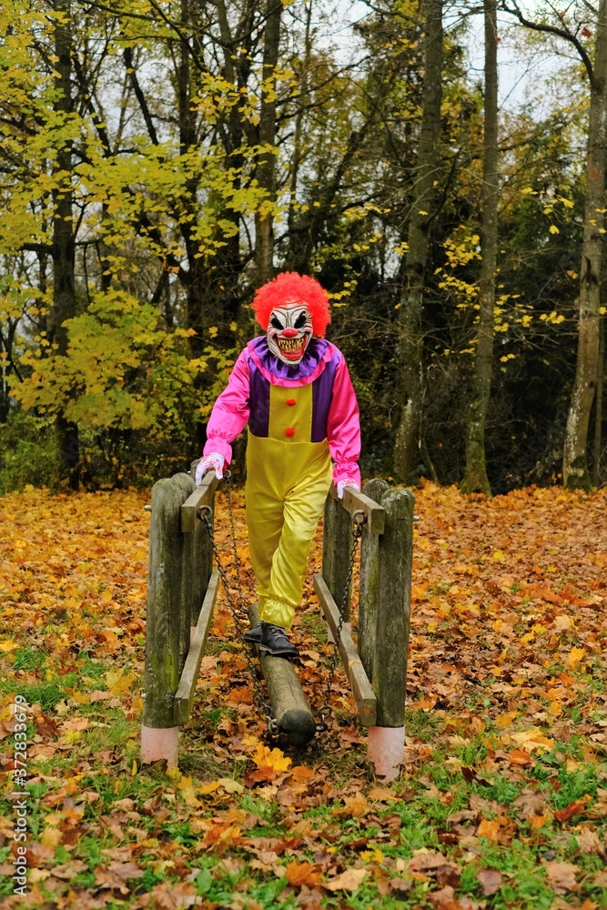 Halloween holiday. Evil clown in overalls and in a scary mask on the playground in the autumn park.Autumn holidays time. halloween mood.Carnival in October Halloween holiday. Evil clown in overalls and in a scary mask on the playground in the autumn park.Autumn holidays time. halloween mood.Carnival in October