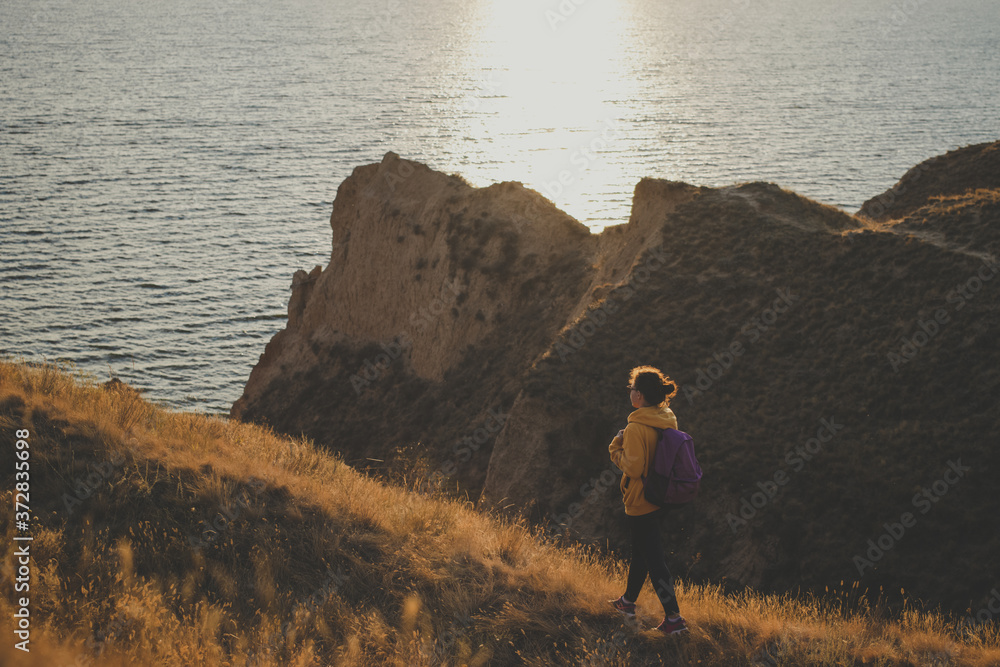 Young woman traveler with backpack in yellow hoodie walking the trail path at mountain peak, hiking to cliff by the sea during sunset, enjoying nature landscape. Soft filter. Adventure concept