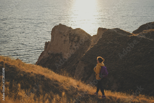 Young woman traveler with backpack in yellow hoodie walking the trail path at mountain peak, hiking to cliff by the sea during sunset, enjoying nature landscape. Soft filter. Adventure concept