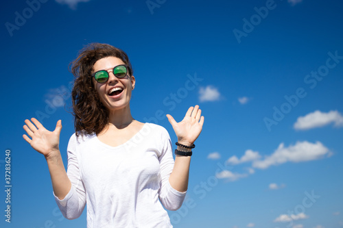 Cheerful young brunette woman in sunglasses saying hi and smiling friendly, gesturing raising hands waiting in greeting or goodbye, joyfully welcome someone, standing outdoors on blue sky background