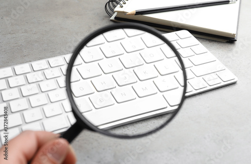Woman holding magnifier glass near keyboard at light grey stone table, closeup. Find keywords concept