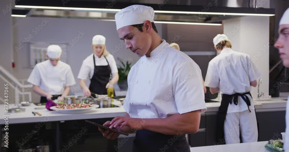 Mixed race male chef wearing chefs whites in a restaurant kitchen using a tablet and looking at came Mixed race male chef wearing chefs whites in a restaurant kitchen using a tablet and looking at came