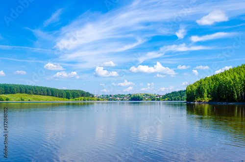 Lake in the forest on a clear summer day. A village on the shore of a reservoir. House. Settlement. Specular reflection in the water. Green foliage of trees. Coast. Blue sky with white clouds on a sun