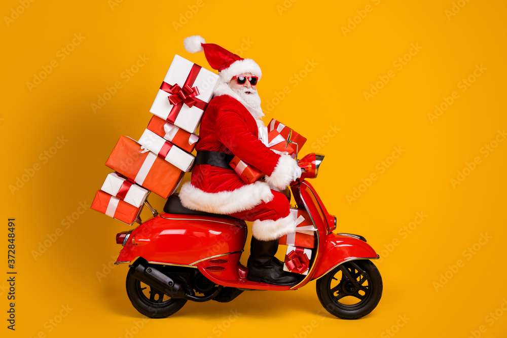 Profile side view of his he nice funny cheery white-haired St Nicholas riding moped hurry up rush delivering bringing pile stack giftboxes isolated bright vivid shine vibrant yellow color background Profile side view of his he nice funny cheery white-haired St Nicholas riding moped hurry up rush delivering bringing pile stack giftboxes isolated bright vivid shine vibrant yellow color background