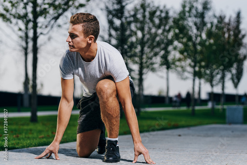 Athletic man in shorts and a t-shirt prepares for jogging in nature in the park near the trees on the street