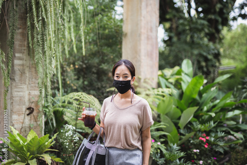 Woman in protective mask hand hold iced coffee walk in greenery park quarantine.