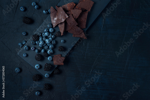 Blueberry and blackberry with chocolate pieces on the black cutting board and dark textured background. Flat lay. Copy space.