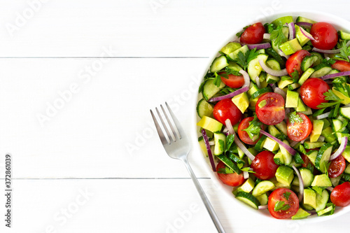 Salad of fresh ripe tomato, onion, avocado, cucumber and parsley and olive oil, salad in a white plate with a fork and towel on a white wooden background
