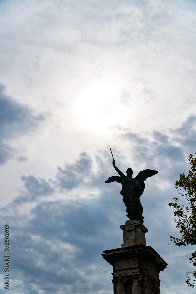 Fototapeta premium Statue of an angel in Rome with sword and clouds background with sun rays as divine light