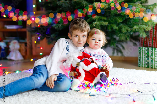 Adorable baby girl and brother holding colorful lights garland and toy Santa Claus in cute hands.