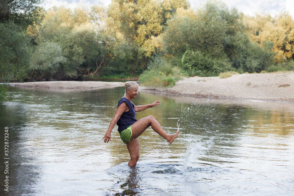Fototapeta premium Senior woman in a shallow river playing with water. Mature woman enyoing in a river, splashing water with a leg.