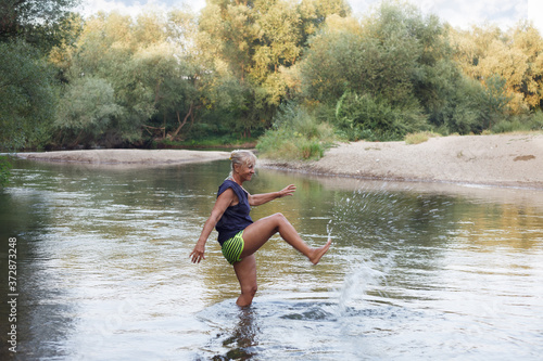 Fototapeta Naklejka Na Ścianę i Meble -  Senior woman in a shallow river playing with water. Mature woman enyoing in a river, splashing water with a leg.
