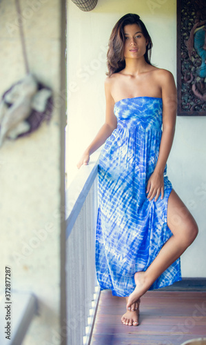 Young beautiful asian brunette woman wearing summer blue dress posing standing on balcony against white wall