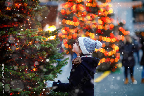 Little cute kid girl having fun on traditional Christmas market during strong snowfall. Happy child enjoying traditional family market in Germany. Schoolgirl standing by illuminated xmas tree. photo