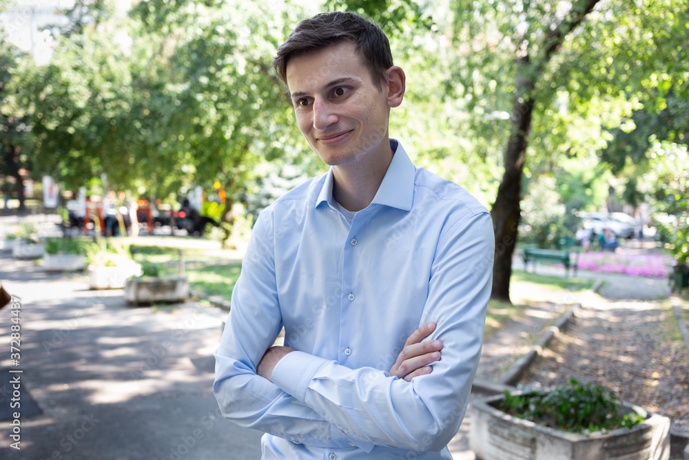 Portrait of young smiled man in a blue shirt on the city with a trees on the background. The man in conversation, listens and speaks. American or European appearance Portrait of young smiled man in a blue shirt on the city with a trees on the background. The man in conversation, listens and speaks. American or European appearance