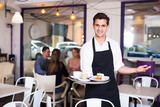 Portrait of happy hospitable waiter inviting to taste coffee and cake