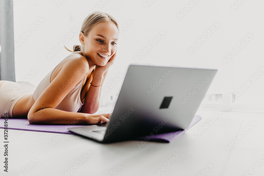 Beautiful young woman watching fitness video tutorials online on laptop while lie down on floor in living room. Beautiful young woman watching fitness video tutorials online on laptop while lie down on floor in living room.
