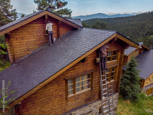 Couple of workers,one on a ladder and one on the roof,working on the facade of a chalet in the mountains