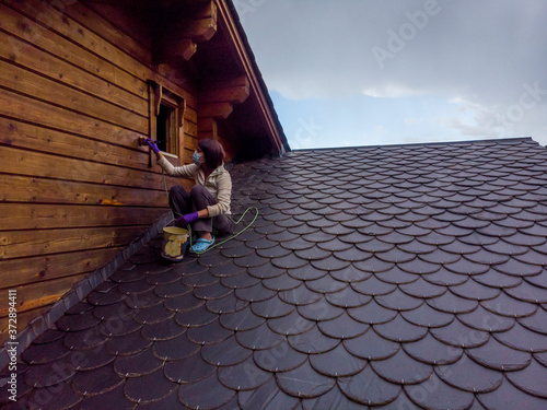 A woman is on a roof of slate tiles varnishing the wall of her chalet