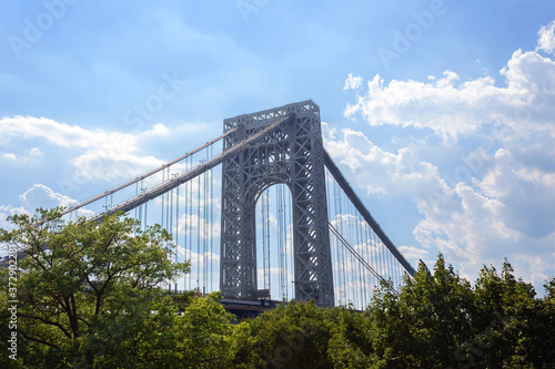 the Manhattan side suspension tower of the George Washington Bridge, a double-decked suspension bridge, above treetops against a bright blue sky with fluffy white clouds with copy space © betzalit