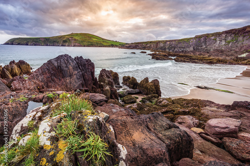 Small sandy hidden beach between cliffs with view on Dingle bay and lighthouse. Dramatic sunset in Dingle peninsula, Co. Kerry, Ireland