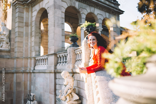 Portrait of cheerful curly brunette woman blogger spending free time outdoors near beautiful city architecture on sunny day, happy charming female traveler holding mobile phone explore locations