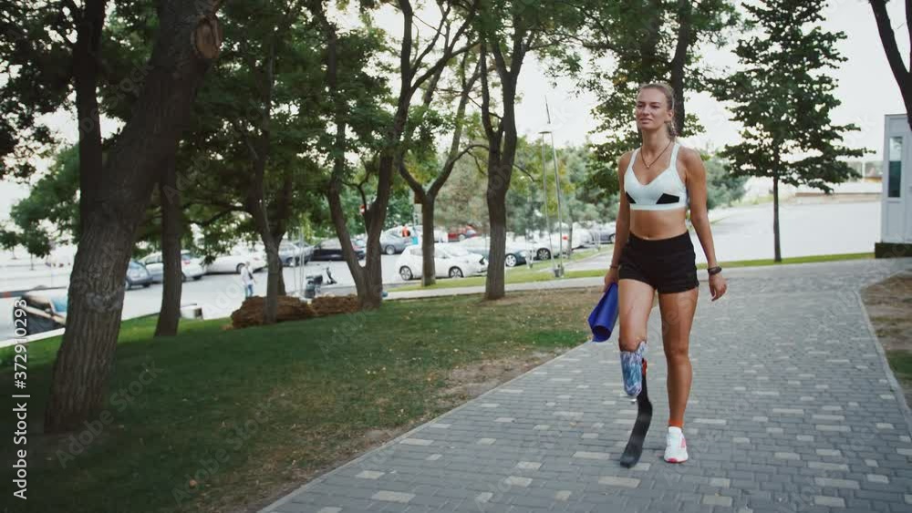 Young girl with leg prosthesis going to training with fitness mat, looking at smartwatch Young girl with leg prosthesis going to training with fitness mat, looking at smartwatch
