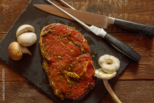 Slice of spiced raw beef with garlic and mushroom on slate plate with meat knife and fork on wooden table