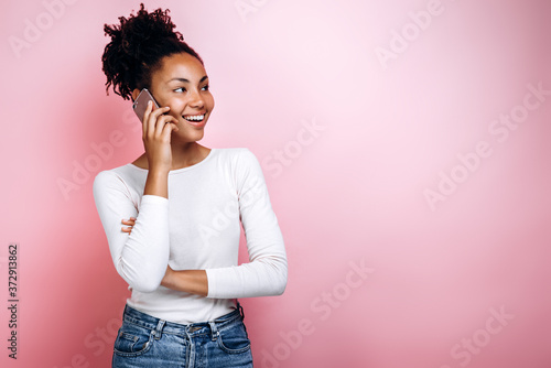 Photo of happy young woman standing isolated over pink background. Looking aside talking by phone.