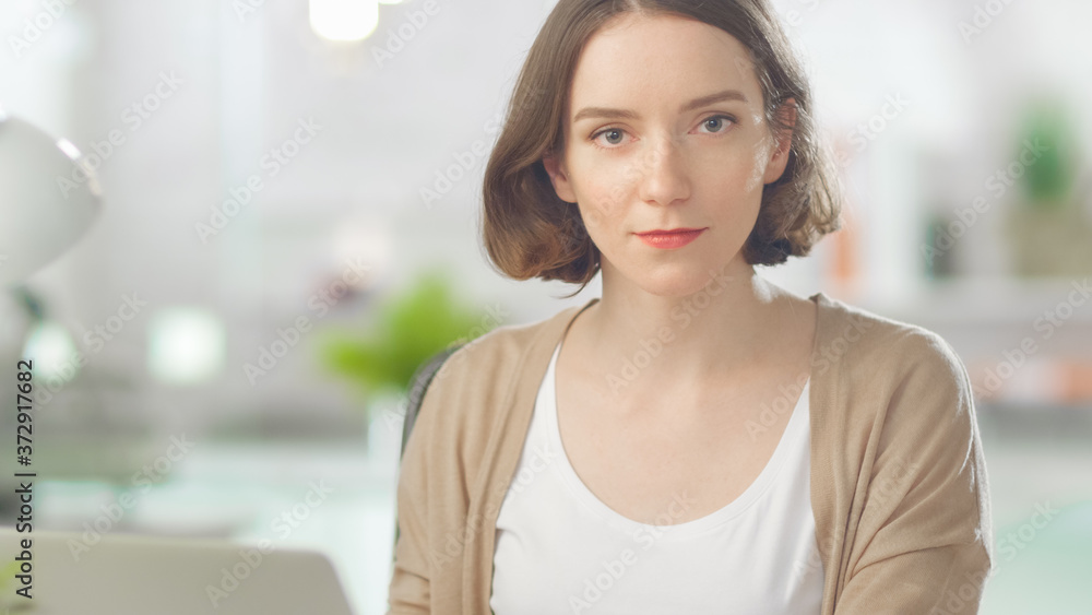 Shot of Focused Young Woman Working on Her Laptop Computer and Looking at Camera. In the Background Blurred Bright Modern Loft Office. Shot of Focused Young Woman Working on Her Laptop Computer and Looking at Camera. In the Background Blurred Bright Modern Loft Office.