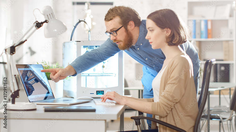 Fototapeta premium Shot of a Young Bearded Male Manager Gives Advice to a Female 3D Engineer, who is using Laptop Computer. Bright Modern Office with Programmable 3D Printer.