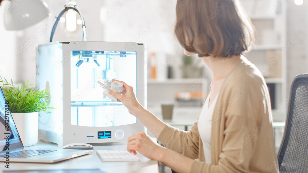 Young Woman Engineer Developer Expects 3D Printed Plastic Part that She Just Made with Her 3D Printer. Bright Modern High Tech Office with Successful People. Young Woman Engineer Developer Expects 3D Printed Plastic Part that She Just Made with Her 3D Printer. Bright Modern High Tech Office with Successful People.
