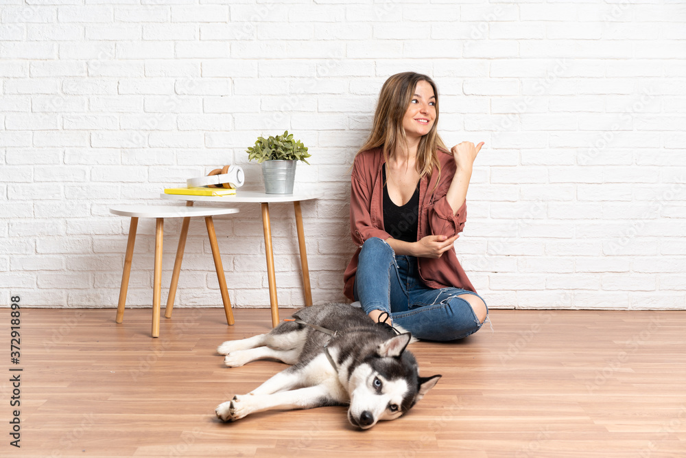 Young girl with her dog sitting in the floor at indoors pointing to the side to present a product