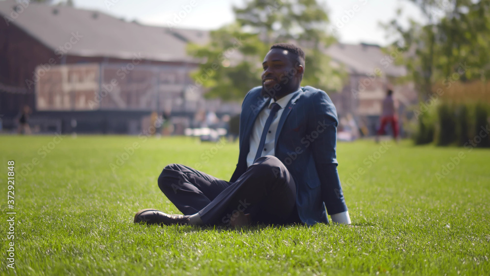 African businessman sitting cross-legged relaxing on green lawn near office center African businessman sitting cross-legged relaxing on green lawn near office center