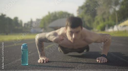 Front view of serious endurant sportsman pushing up on athletic field in the morning. Portrait of confident young Caucasian tattooed man training in sunlight outdoors. Masculinity concept. photo