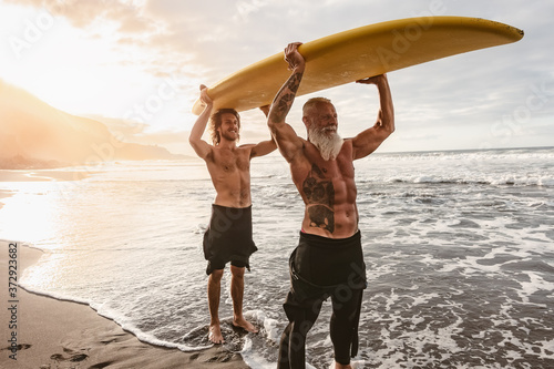 Happy friends with different age surfing together on tropical ocean - Sporty people having fun during vacation surf day - Elderly and youth people and extreme sport lifestyle concept