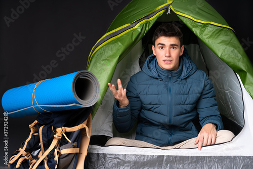 Teenager caucasian man inside a camping green tent isolated on black background making doubts gesture