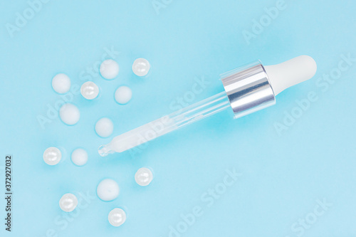 Cosmetic pipette with serum on blue background. Around - drops of serum and pearl. Stylish concept of organic essences, beauty and health products. Copy space, minimalism, flat lay. Modern apothecary.