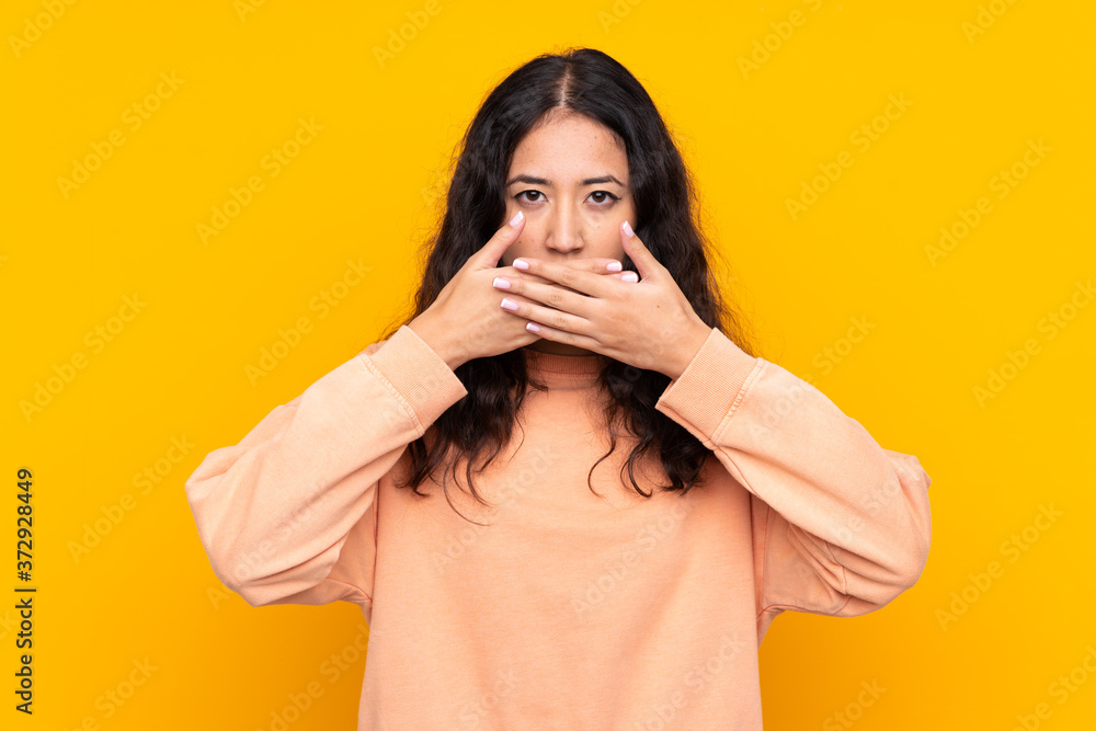 Spanish Chinese woman over isolated yellow background covering mouth with hands Spanish Chinese woman over isolated yellow background covering mouth with hands
