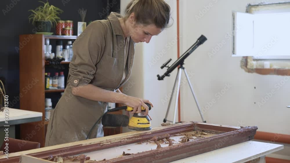 Young woman grinds the wooden door, restoration works. Female and a yellow grinder, polishing old door in workshop handheld shot, renovation of old-fashioned product Young woman grinds the wooden door, restoration works. Female and a yellow grinder, polishing old door in workshop handheld shot, renovation of old-fashioned product