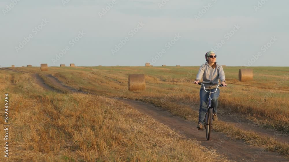 A bicyclist girl in a headscarf rides a Bicycle on a rural road full of twisted straw bales in a field. A bicyclist girl in a headscarf rides a Bicycle on a rural road full of twisted straw bales in a field.