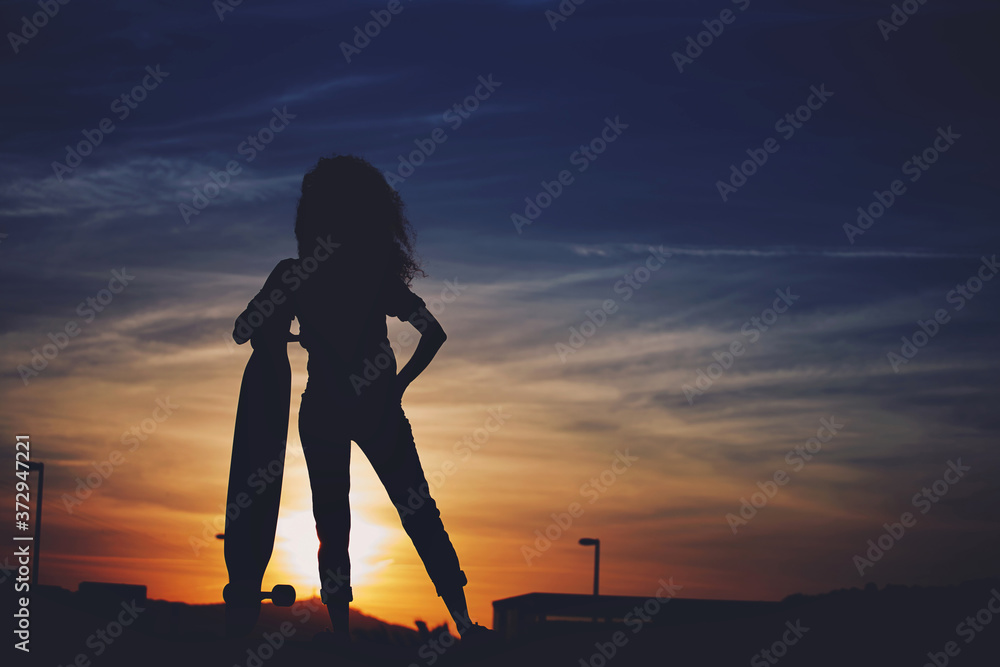 Silhouette of a skater girl with perfect figure stands against dramatic sunset background with copy space area for text message or content, female enjoying the heavenly scenery with a board to ride Silhouette of a skater girl with perfect figure stands against dramatic sunset background with copy space area for text message or content, female enjoying the heavenly scenery with a board to ride