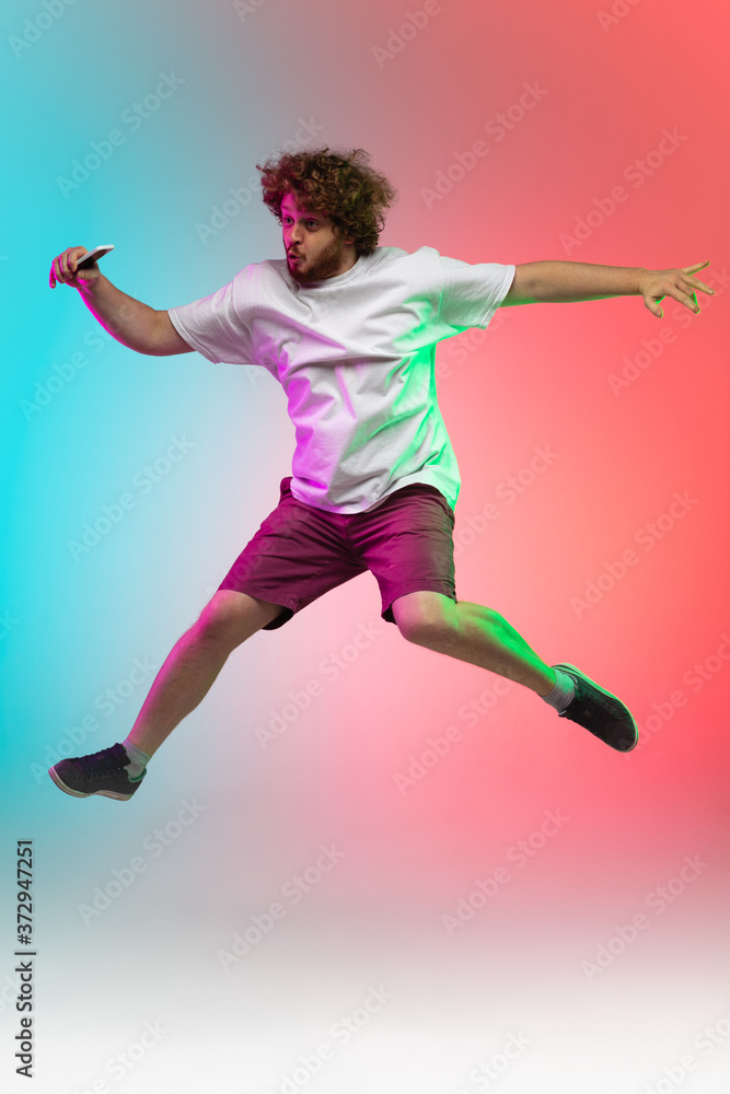 Phone scrolling. Caucasian young man's portrait on gradient studio background in neon. Beautiful male model in casual style jumping high. Concept of human emotions, facial expression, youth, sales, ad