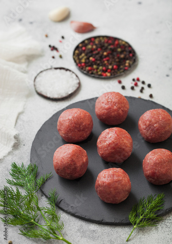 Fresh beef raw meatballs on round stone board with pepper, salt and garlic on light background with dill and towel.