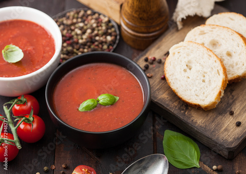 Ceramic bowl plates of creamy tomato soup with spoon, pepper and kitchen cloth on wooden background with raw tomatoes and bread.