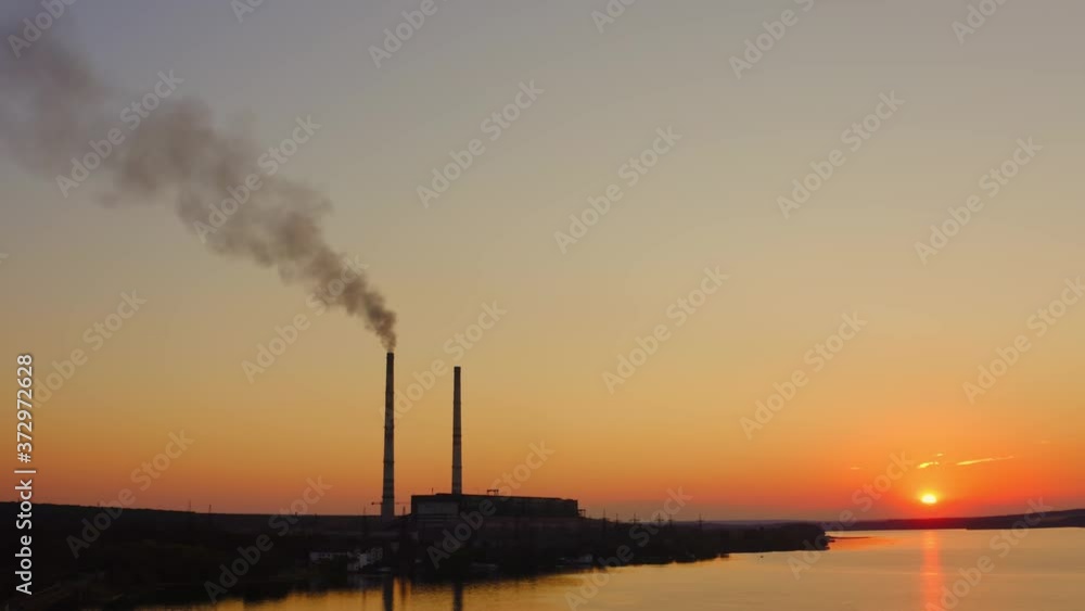 Plant with smoke against orange sky. Industry among nature at sunset. Harmful smoke released from factory in the air.