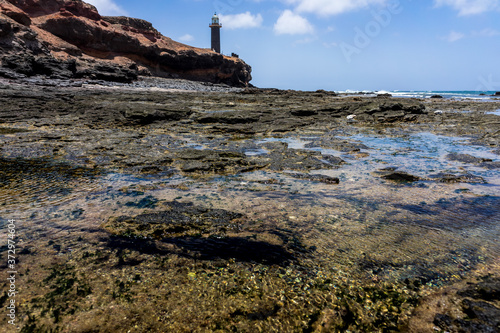 Rocky Atlantic coastline at low tide. In the background, the Punta Jandia lighthouse. Fuerteventura. Canary Islands. Spain.