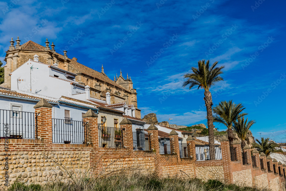 Brick wall with pillars and wrought iron bars in a street on a hill with palm trees with houses and the Royal Collegiate Church of Santa María la Mayor in the background, sunny day in Antequera, Spain
